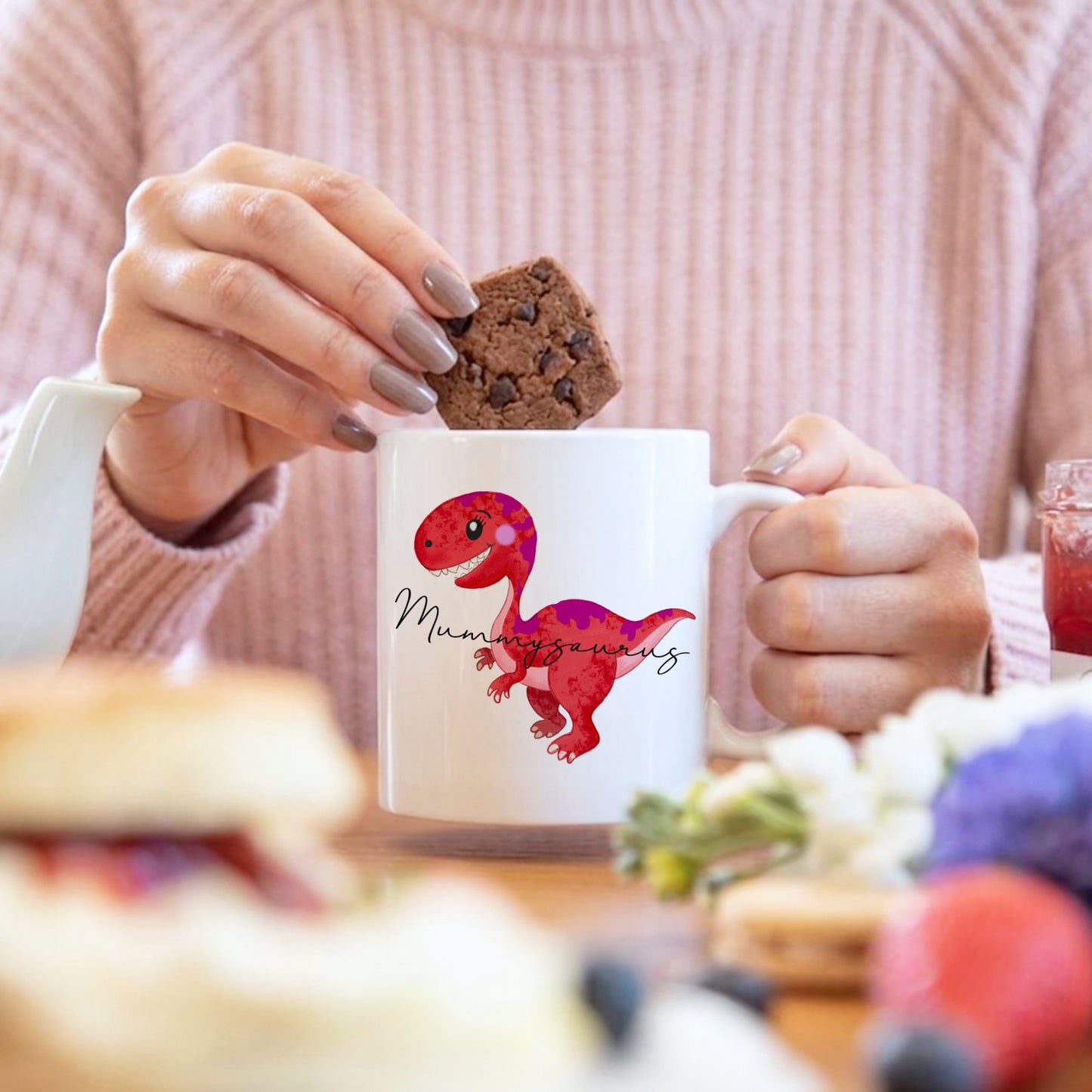Person holding a mug with a dinosaur design and a cookie, blurred background