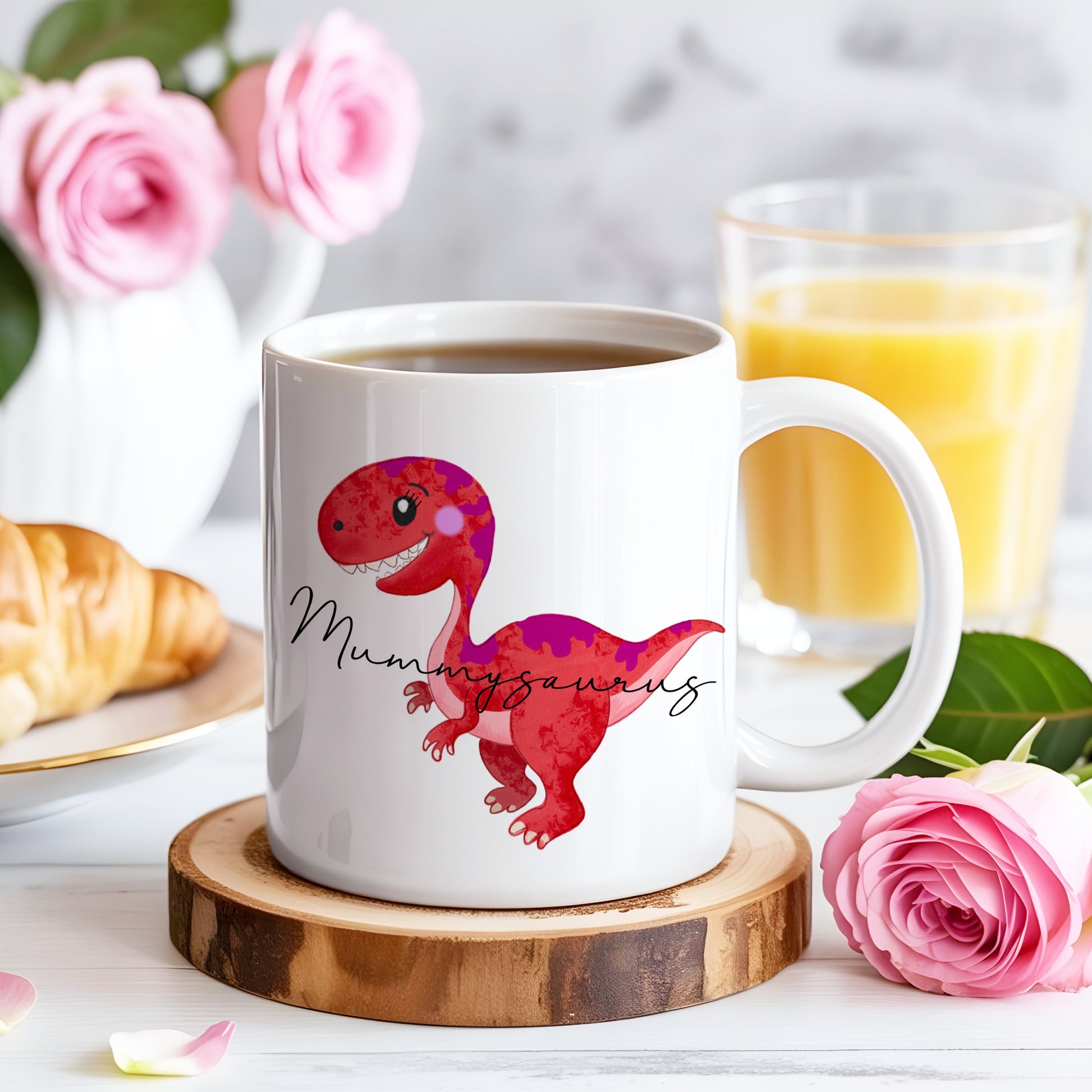 White mug with a red dinosaur design and 'Mummyaurus' text on a wooden coaster, surrounded by pink roses and a glass of orange juice.