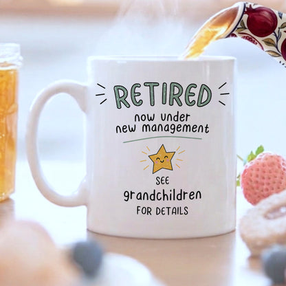 A white ceramic mug with a humorous retirement quote printed on it, shown in a lifestyle setting with tea being poured into it from a floral teapot, surrounded by strawberries and a jar of honey.