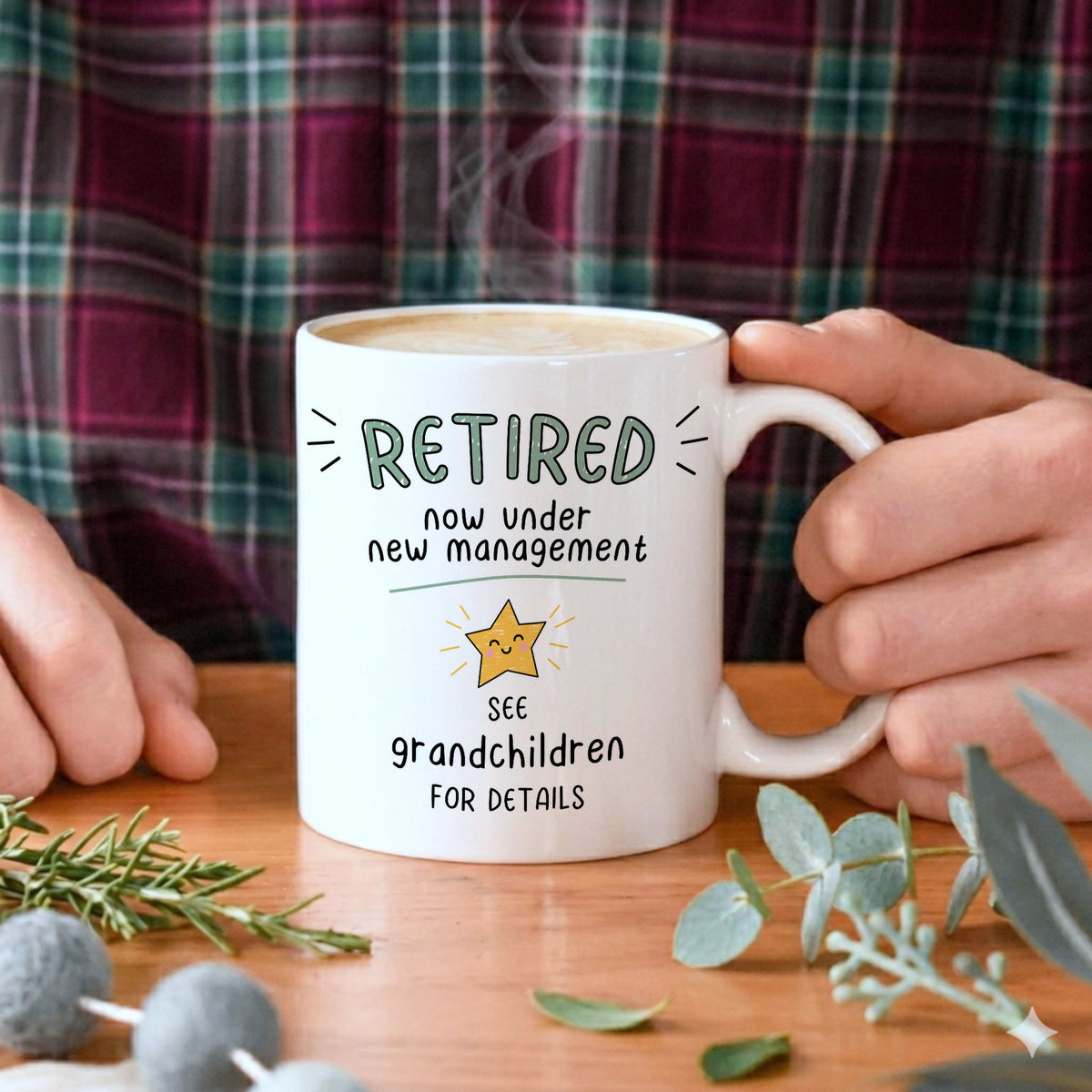 A white ceramic mug with a humorous retirement message printed on it, held by hands against a background of a person wearing a green and red plaid shirt, with eucalyptus leaves and rosemary on a wooden table.