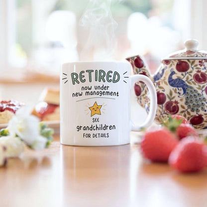 A white ceramic mug with a humorous retirement quote printed on it, styled on a wooden table with a teapot and strawberries in a warm, blurred kitchen background.