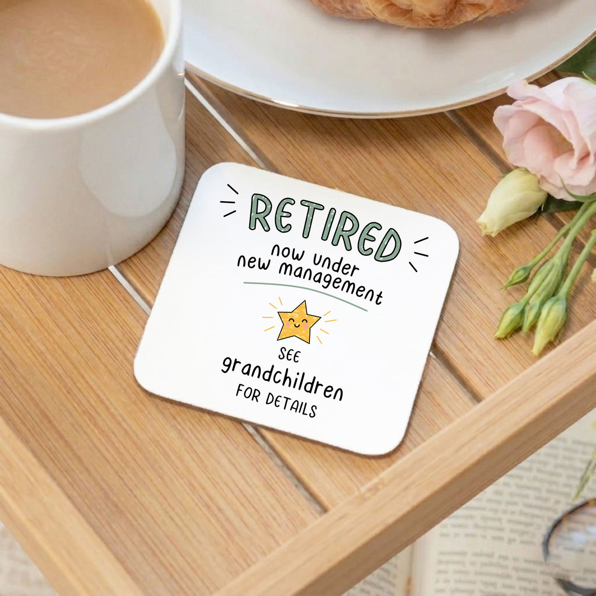 A square white coaster with rounded corners sits on a wooden tray. The coaster has a printed design that reads 'RETIRED now under new management' with a small star icon and 'SEE grandchildren FOR DETAILS'.