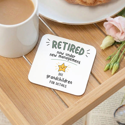 A square white coaster with rounded corners sits on a wooden tray. The coaster has a printed design that reads 'RETIRED now under new management' with a small star icon and 'SEE grandchildren FOR DETAILS'.
