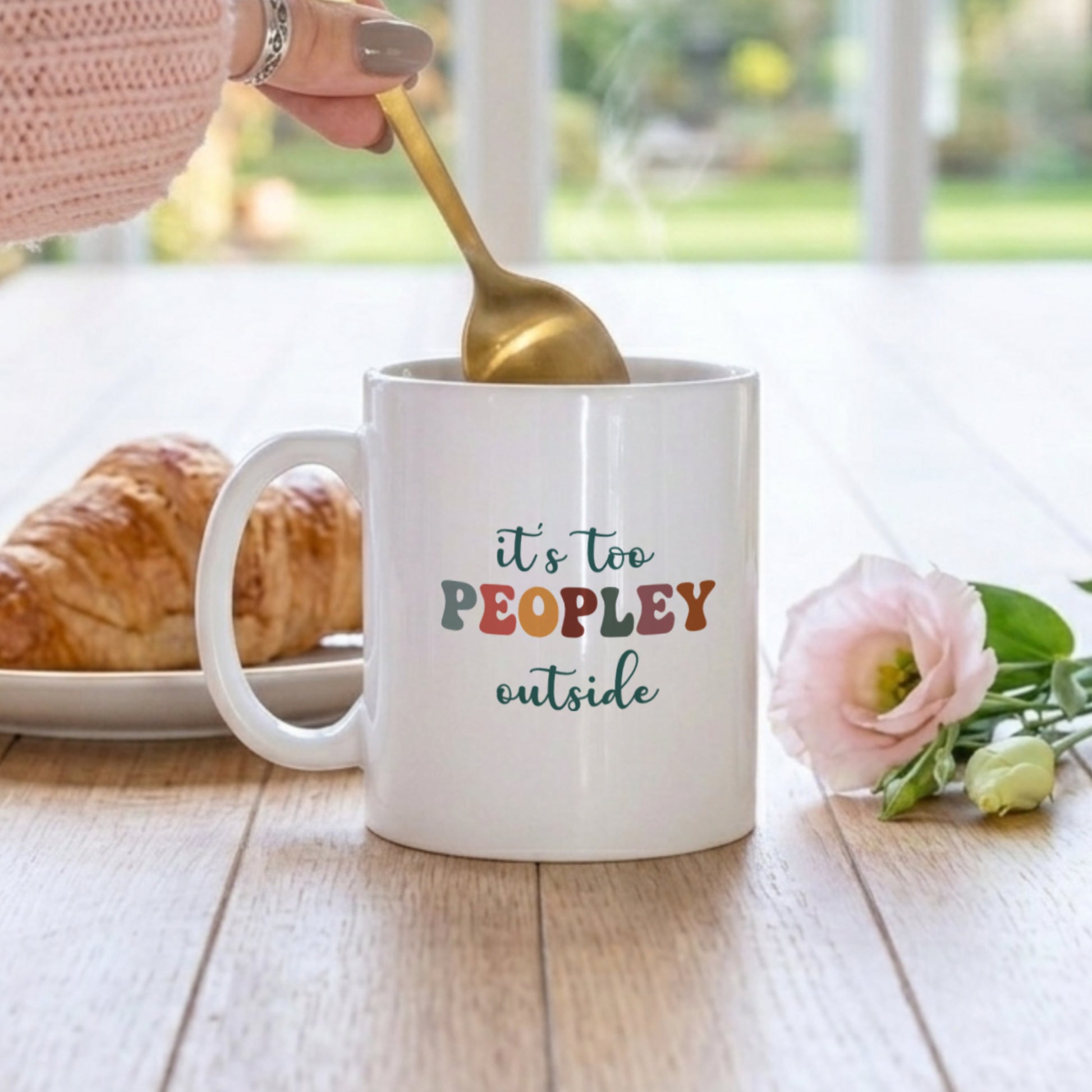 White mug with colorful text 'it's too peopley outside' on a wooden table with a croissant and flowers.