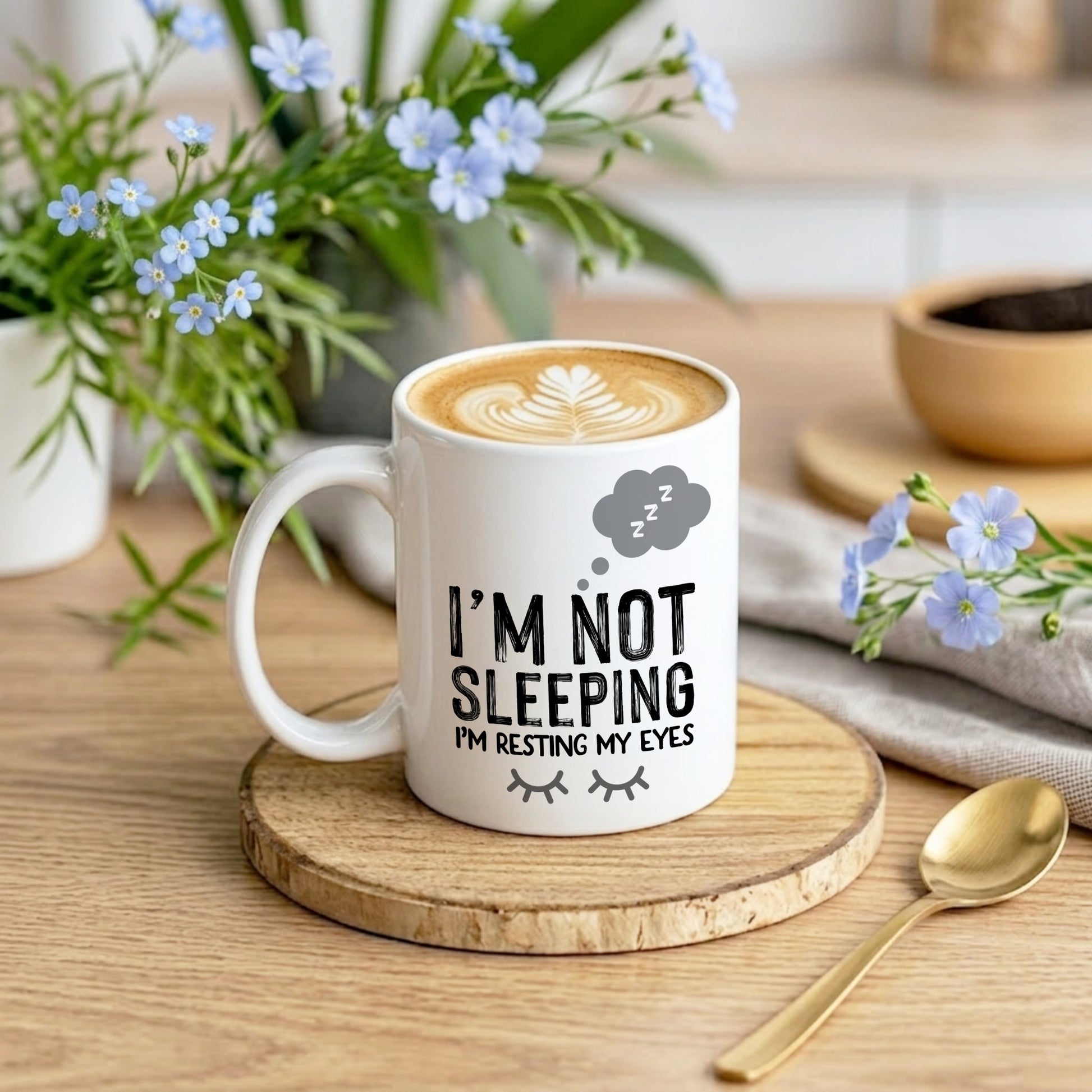 Mug with 'I'm not sleeping, I'm resting my eyes' text on a wooden coaster with flowers and a spoon in the background.