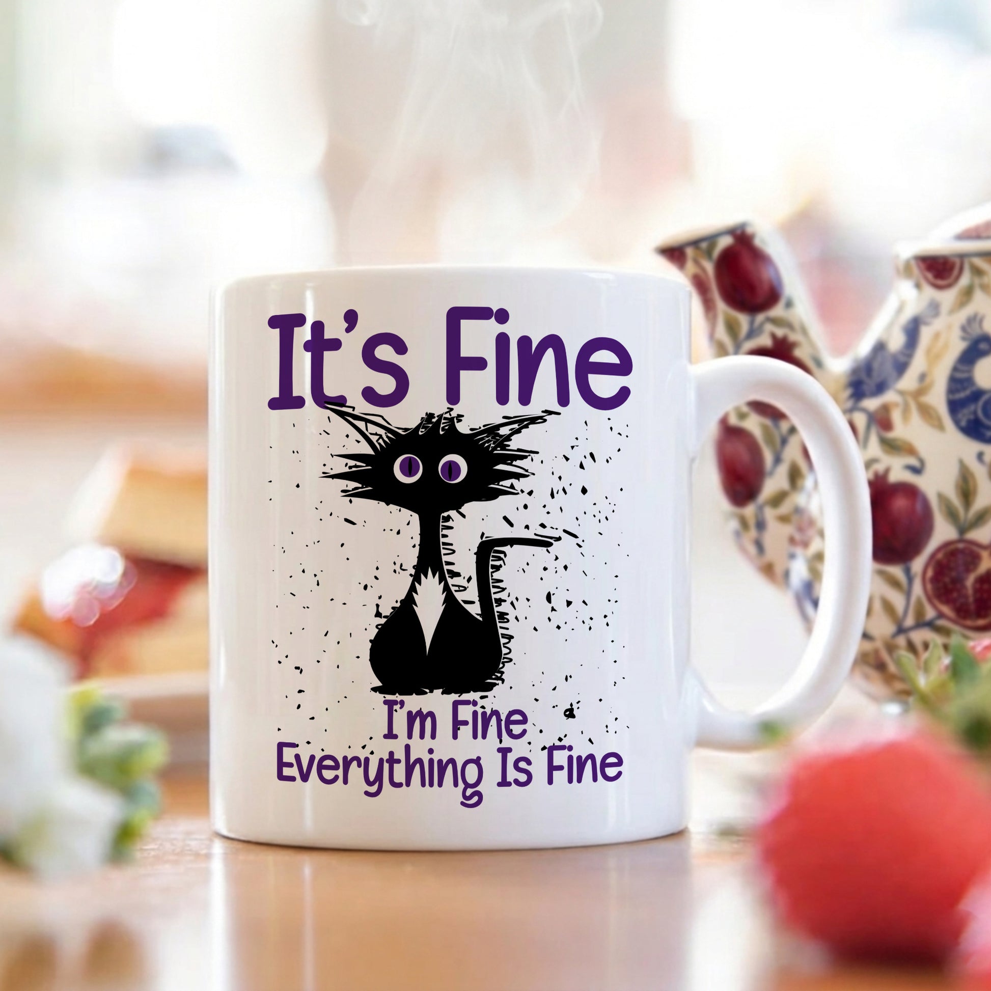 White mug with a black cat illustration and text 'It's Fine, I'm Fine, Everything Is Fine' on a blurred background of a kitchen.