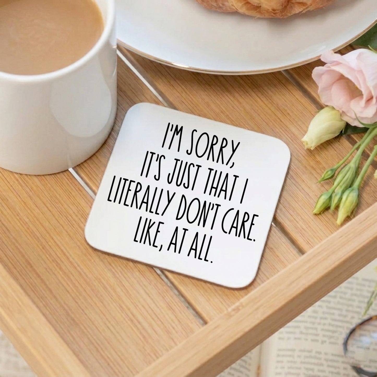 A white square coaster on a breakfast tray with the text "I'm sorry, it's just that I don't care. Like, at all". With a cup of tea, croissant and pink rose 
