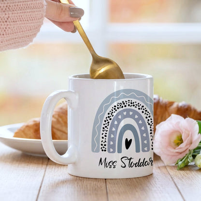 Personalised teacher mug with a blue boho rainbow and heart design, customised with 'Miss Stoddart', being stirred with a gold spoon alongside a croissant and pink flower