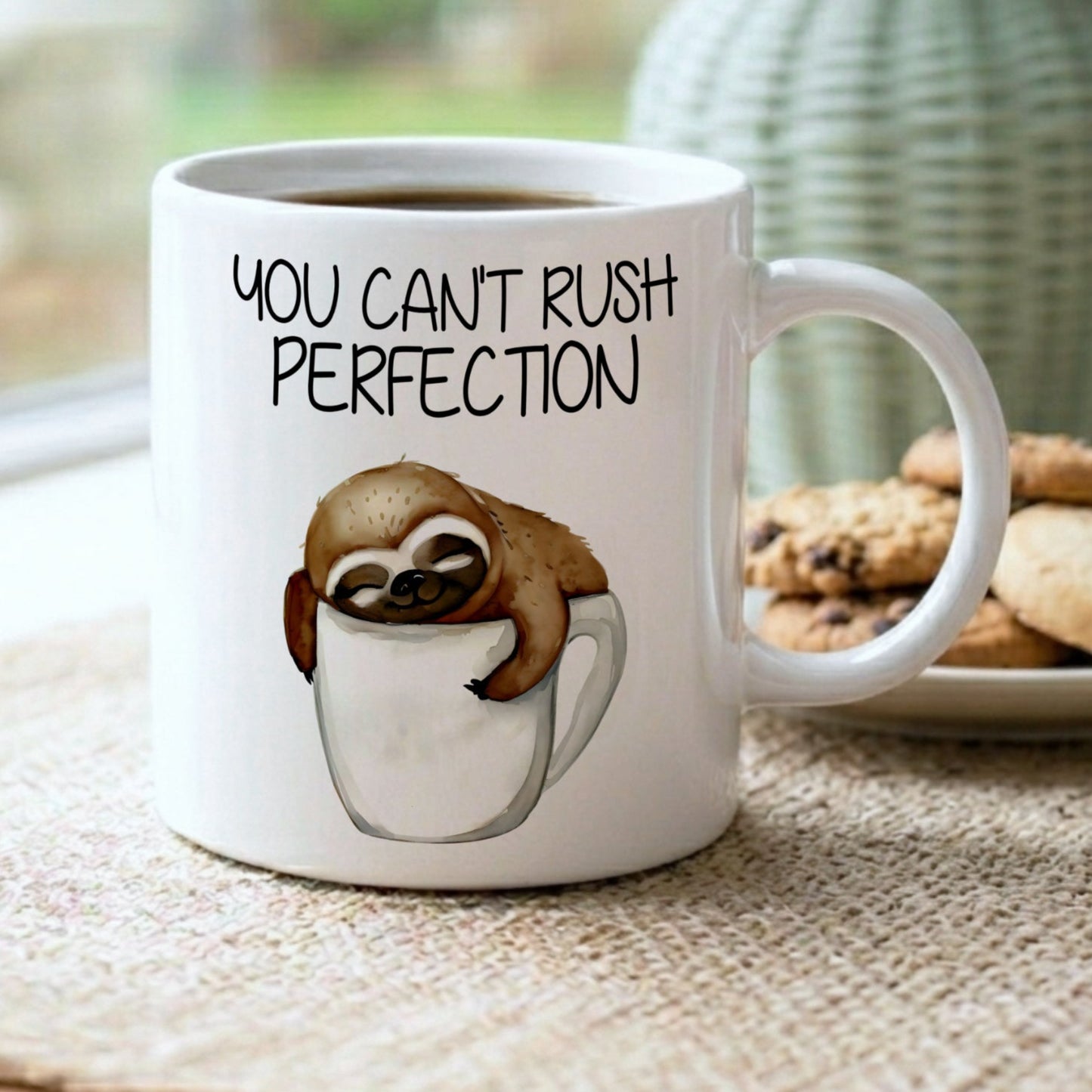 Mug with sloth illustration and 'You can't rush perfection' text on a table with cookies and a vase.