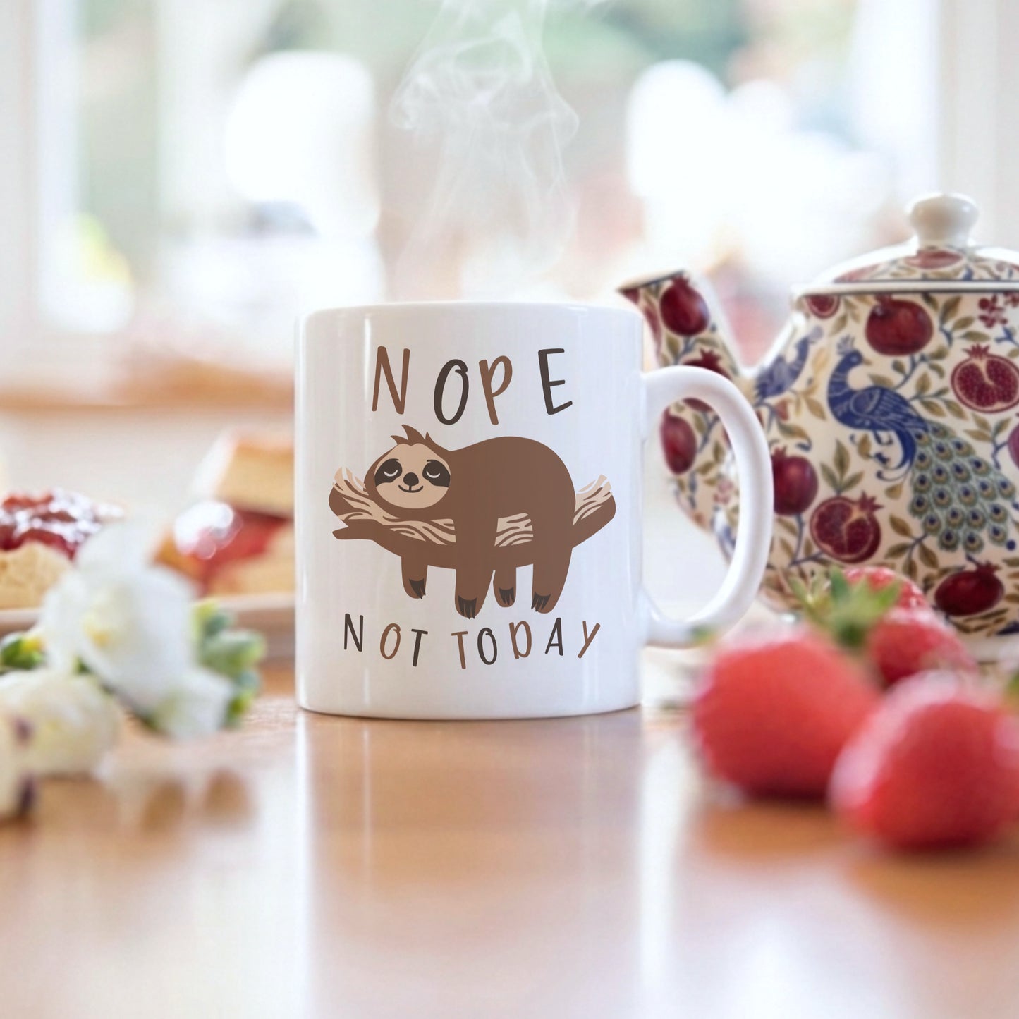 'Nope Not Today' sloth mug displayed on a kitchen table alongside a floral teapot, fresh strawberries, and white flowers