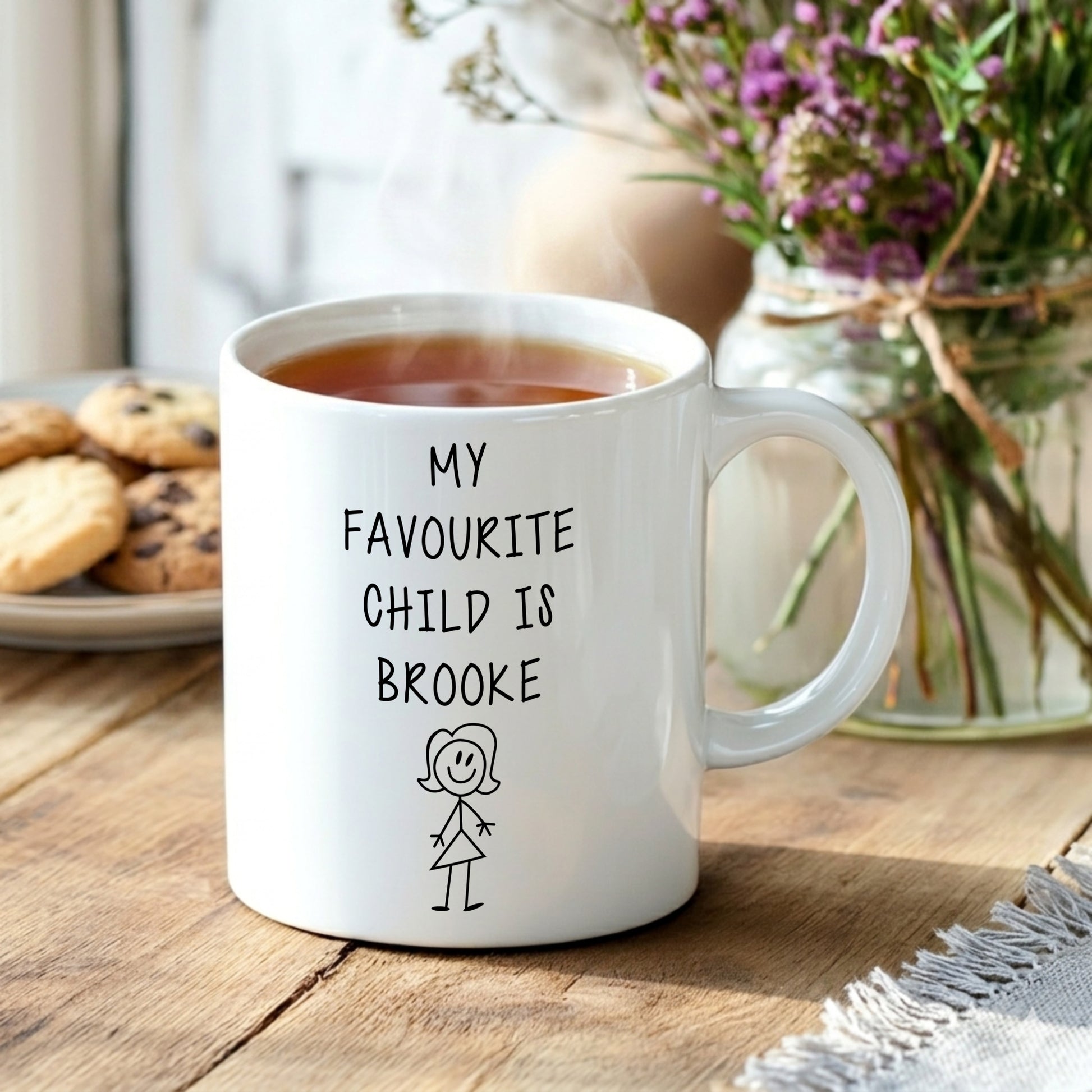 White mug with 'My favourite child is Brooke' text on a wooden table with cookies and flowers.