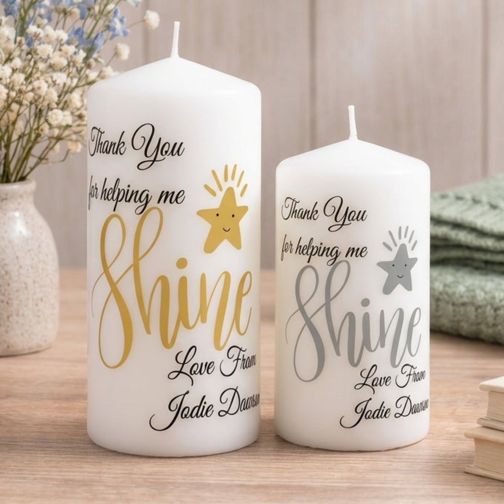 Two white pillar candles with personalised messages printed on them, one smaller and one larger, both with the same design and colour scheme, placed on a red surface.