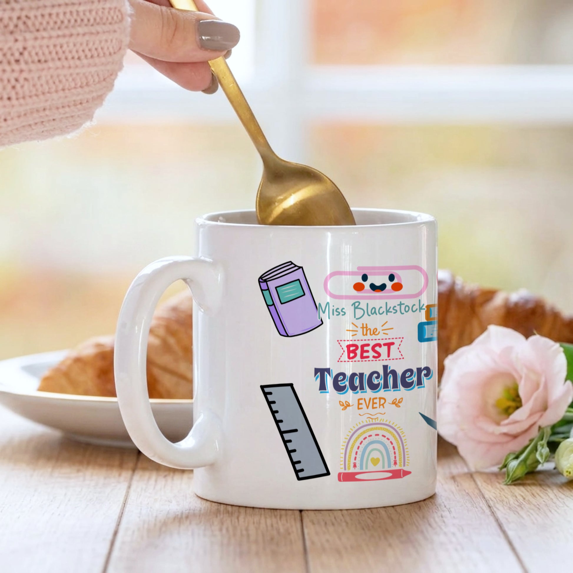 Mug with 'The Best Teacher Ever' text and graphics, being stirred with a spoon.