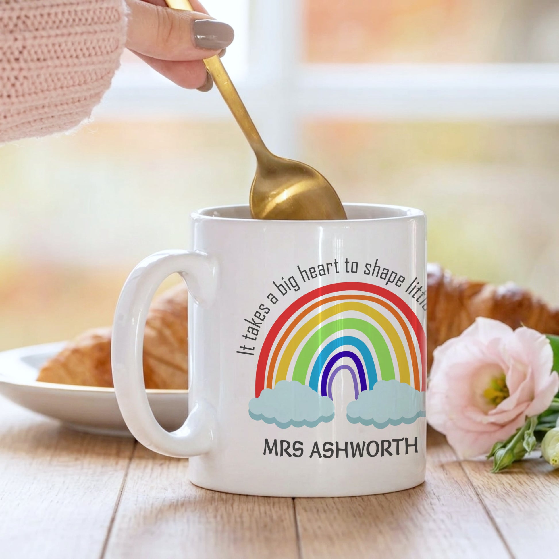White mug with rainbow design and text, held by a hand with a gold spoon, on a wooden table.