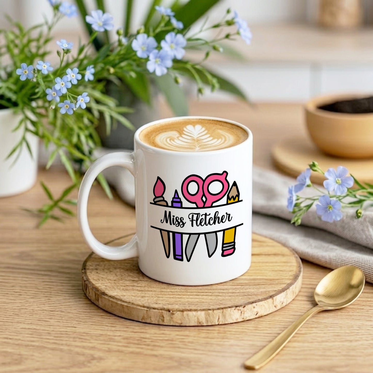 White mug with a personalized design on a wooden coaster, surrounded by flowers and kitchen items.