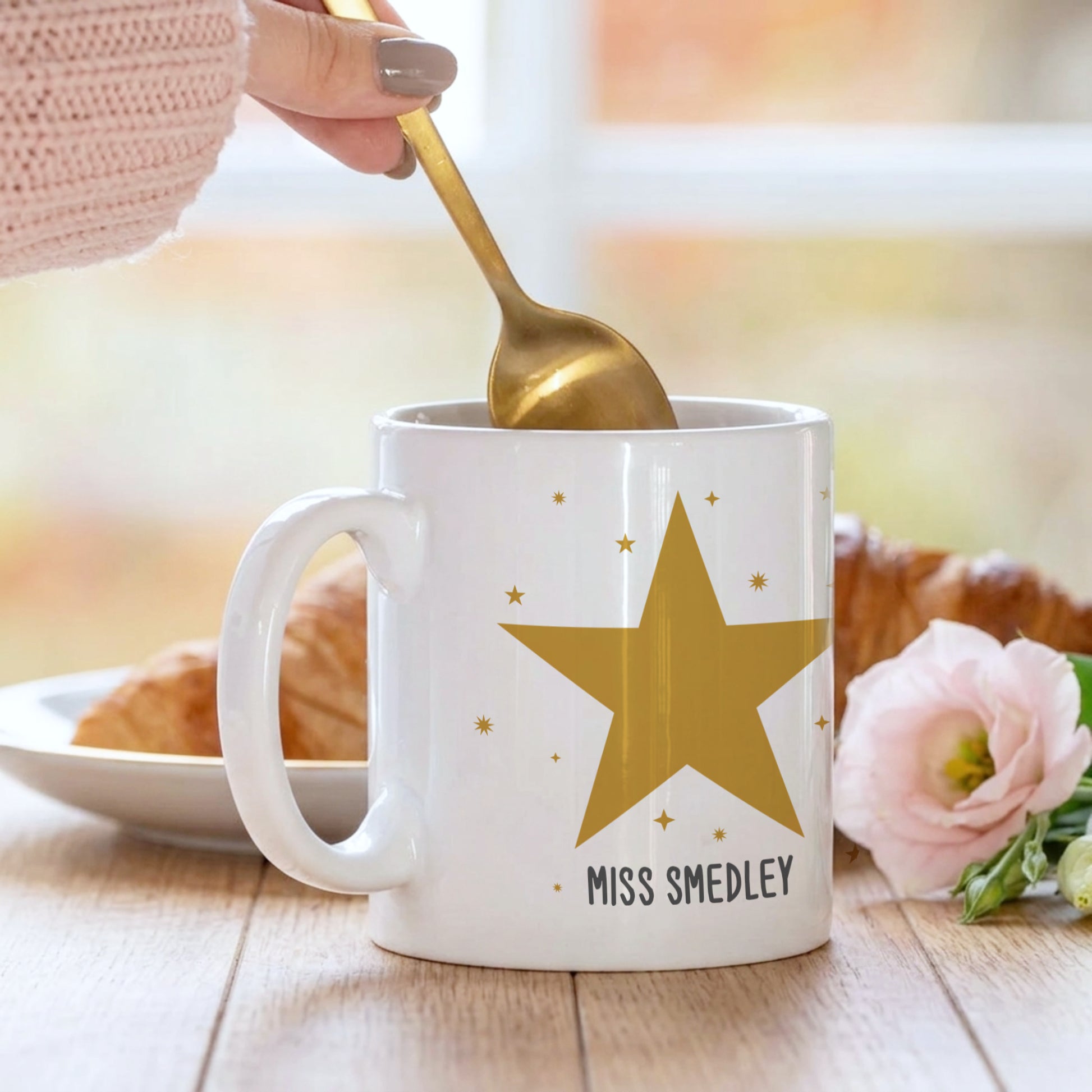 White mug with a gold star and 'MISS SMEDLEY' text, being stirred with a gold spoon.
