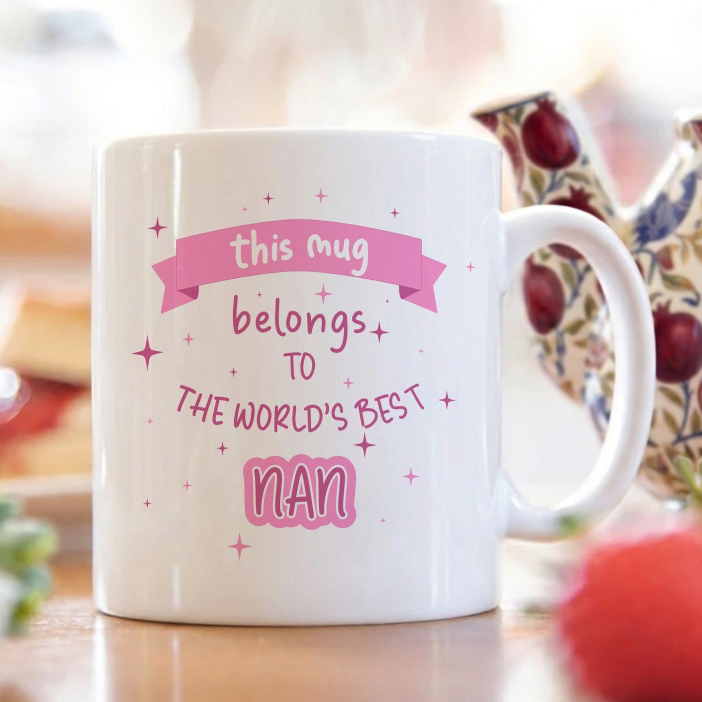 A white ceramic mug with pink text reading 'this mug belongs to THE WORLD'S BEST nan' decorated with small pink stars, shown on a table with a teapot and strawberries in a blurred kitchen setting.