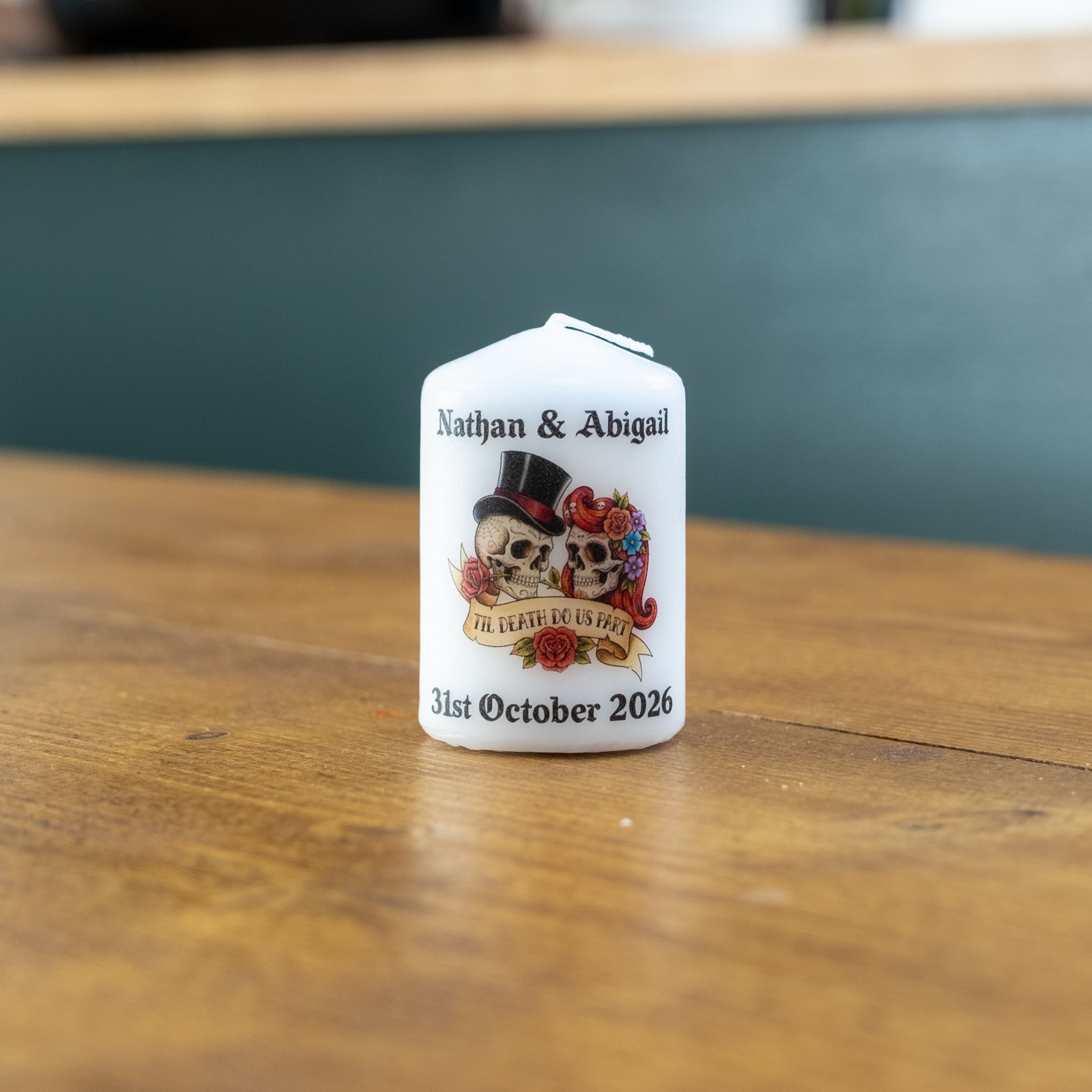 A small white personalised candle standing on a wooden table, printed with the names Nathan & Abigail, a skull-and-rosette illustration, and the date 31st October 2026.