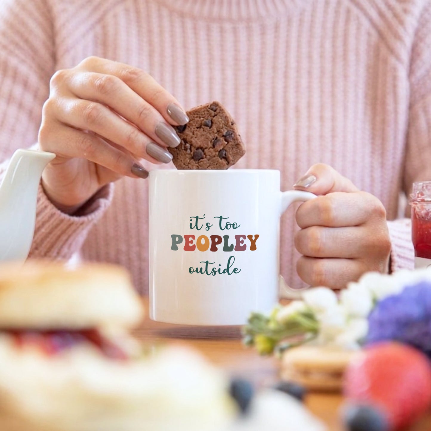 Person holding a mug with 'it's too PEOPLEY outside' text and a cookie.