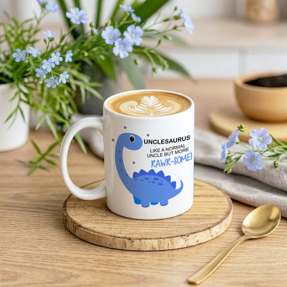 A white ceramic coffee mug with a blue dinosaur illustration and the text 'UNCLESAURUS LIKE A NORMAL UNCLE BUT MORE RAWR-SOME!' sits on a wooden coaster on a wooden table. The mug is styled in a cafe-like setting with a latte, flowers, a spoon, and a bowl in the background.
