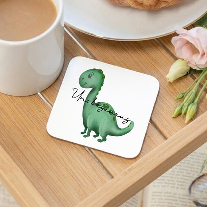 A square white coaster with rounded corners sits on a wooden tray, printed with a green cartoon dinosaur and the words 'Unclesaurus.' A white mug, plate, pastry, and pink flower are visible around it in a styled tabletop setting.