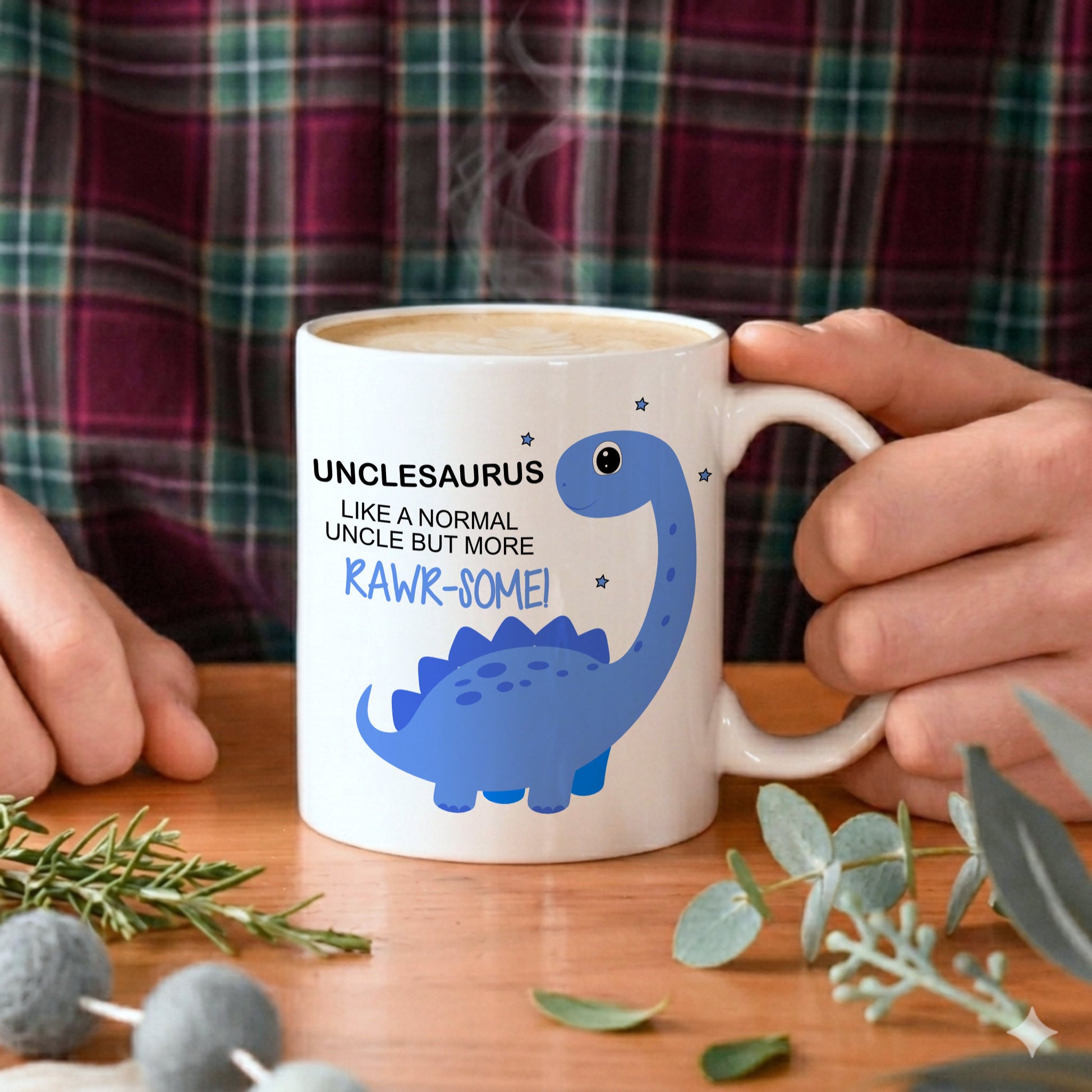 A white ceramic mug with a blue dinosaur graphic and the text 'UNCLESAURUS LIKE A NORMAL UNCLE BUT MORE RAWR-SOME!' is held by hands on a wooden table, with a plaid shirt visible in the background.