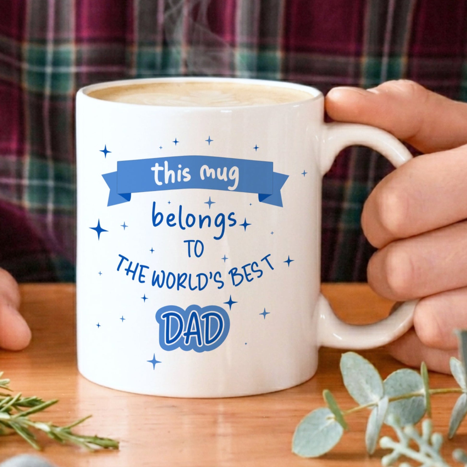 Person holding a mug with 'world's best dad' text on a wooden table with plants.
