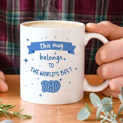 Person holding a mug with 'world's best dad' text on a wooden table with plants.