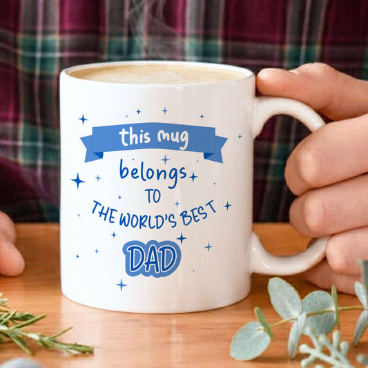 Person holding a mug with 'world's best dad' text on a wooden table with plants.