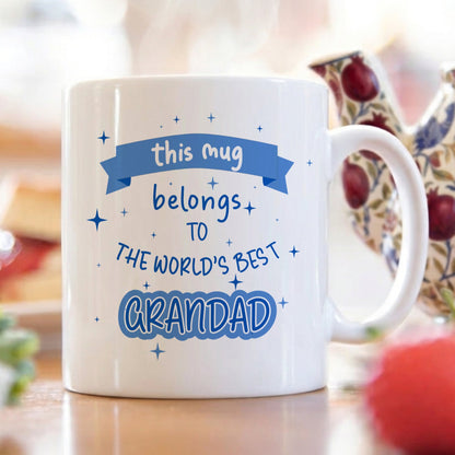 White mug with "this mug belongs to the World's best Grandad" text in blue on a breakfast table with a burred background