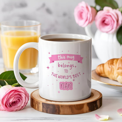 A white ceramic mug filled with coffee sits on a wooden coaster in a breakfast setting. The mug has a pink graphic that reads 'this mug belongs to THE WORLD'S BEST NAN'.