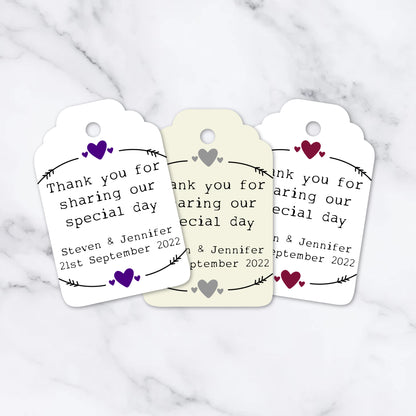 Three personalized heart-shaped wedding tags with white card stock, featuring printed names and a heart design, and a ribbon attachment at the top.